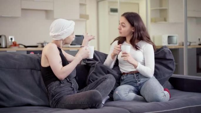 两个年轻漂亮的积极女人坐在沙发上聊天，喝茶或咖啡。快乐放松的高加索朋友在室内休息闲聊和微笑的肖像。友
