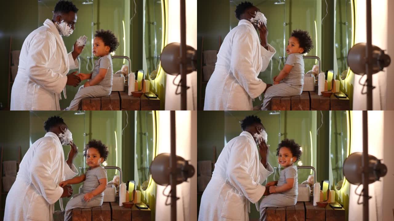 顽皮的年轻父亲用剃须泡沫与坐在浴室里的可爱的蹒跚学步的儿子交谈。积极快乐的非洲裔美国人早上刮胡子和孩