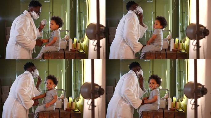 顽皮的年轻父亲用剃须泡沫与坐在浴室里的可爱的蹒跚学步的儿子交谈。积极快乐的非洲裔美国人早上刮胡子和孩