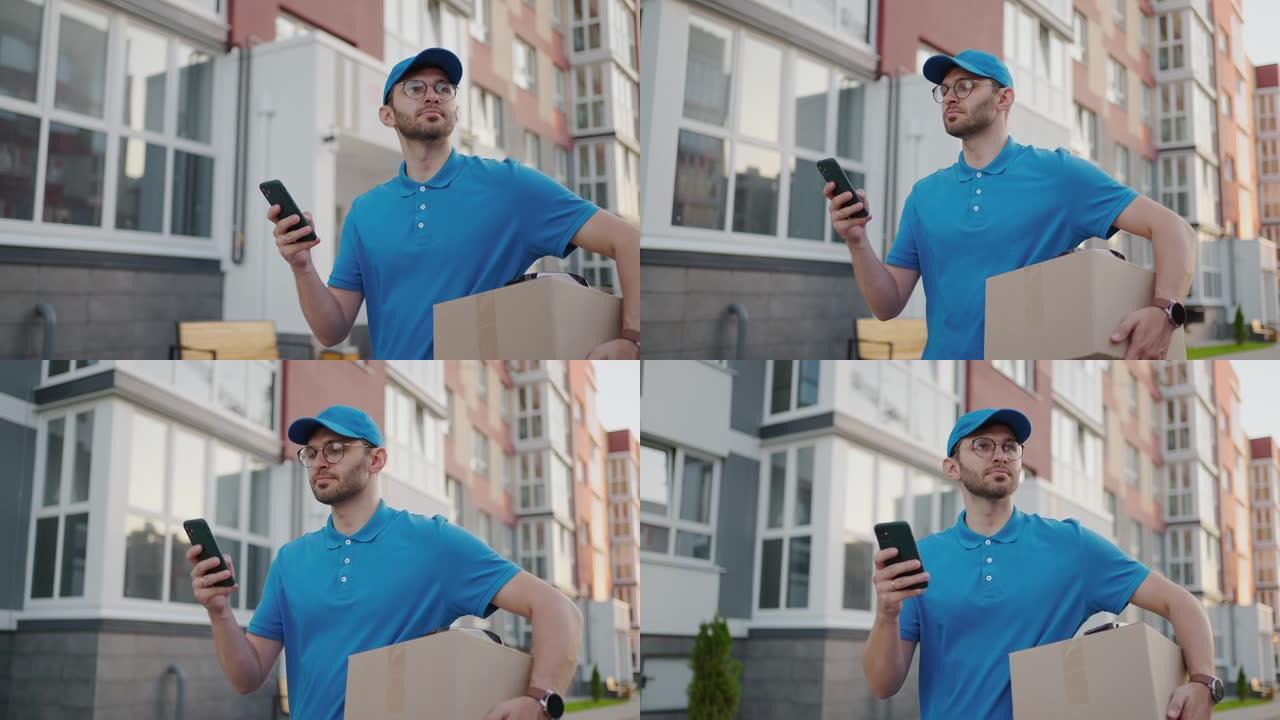 高加索年轻漂亮的男人，穿着蓝色帽子的送货员走在街上，拿着纸箱，同时使用智能手机，寻找路线。男性快递员