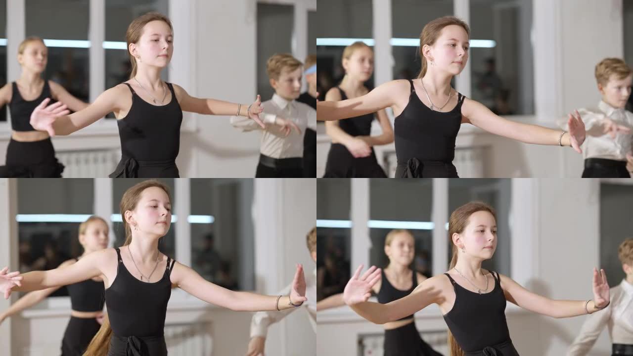 自信的黑发苗条女孩在舞蹈工作室慢动作跳拉丁舞的肖像。集中的高加索儿童与背景朋友排练舞厅艺术。艺术概念