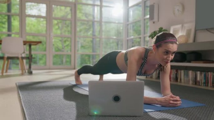 女运动员用笔记本电脑在瑜伽垫上做木板。家庭锻炼