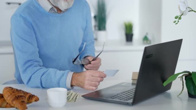 面目全非的大胡子老人在笔记本电脑键盘上打字，拿着名片，摘下眼镜。灰发的高加索退休人员在家室内在线留言