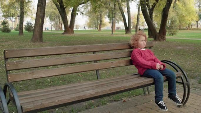 孤独的红发孩子坐在公园的长凳上，被遗弃的孩子感到沮丧