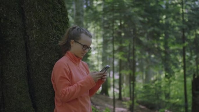使用智能手机的年轻女子望着森林拍照，靠在树上
