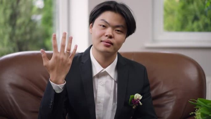 快乐的新婚亚洲男子露出手指，带着戒指微笑看着相机。欢快的新郎坐在舒适的扶手椅上在室内摆姿势的肖像。幸