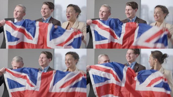 微笑着兴奋的男人和女人拿着英国国旗跳跃。积极自信的成功同事在室内办公室欢欣鼓舞。商业成功和快乐概念。