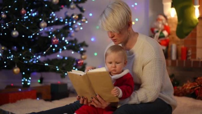 古玩漂亮的女婴在除夕夜在家和年轻漂亮的女人一起读书。可爱的高加索蹒跚学步的孩子在圣诞节和阿姨在室内享