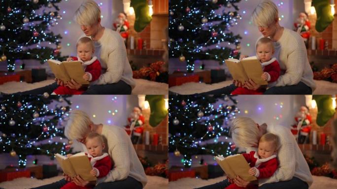古玩漂亮的女婴在除夕夜在家和年轻漂亮的女人一起读书。可爱的高加索蹒跚学步的孩子在圣诞节和阿姨在室内享