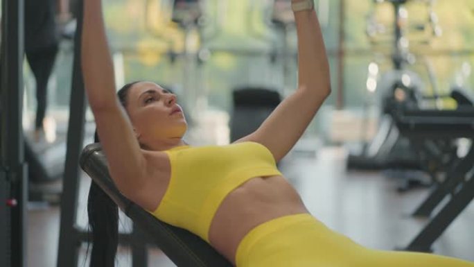 英俊的运动女子躺在健身房里通过繁殖哑铃来训练胸肌。肌肉发达的女运动员用哑铃锻炼，举重