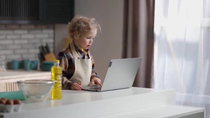 有趣的小厨师穿着围裙正在用笔记本电脑上网，小男孩在家里的厨房里