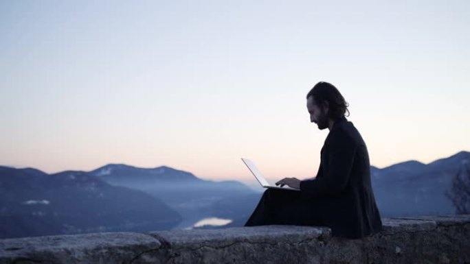 商人坐在笔记本电脑上的边缘工作，日落时可以看到下面的山和湖