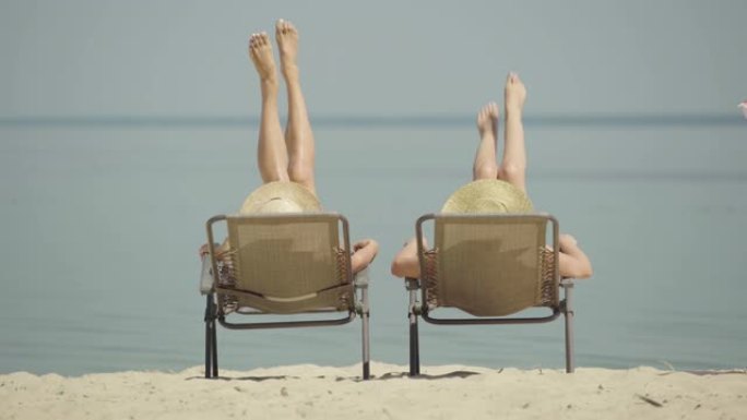 在避暑胜地，轻松的高加索女性摇晃着晒黑的纤细的腿。面目全非的年轻游客躺在日光浴躺椅上晒日光浴。快乐富