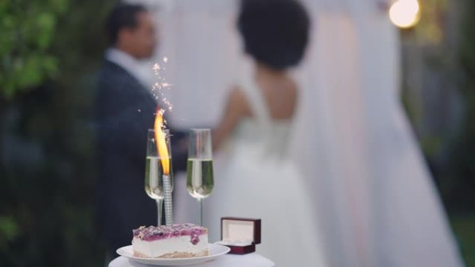 婚礼蛋糕中的特写婚礼烟火和两个香槟酒杯，背景是幸福的情侣牵手。日落时分，快乐模糊的非裔美国新婚夫妇在