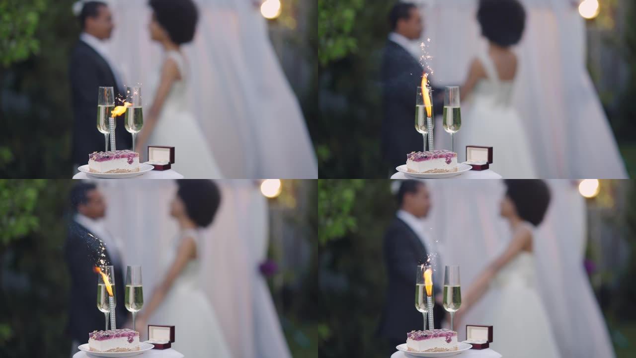 婚礼蛋糕中的特写婚礼烟火和两个香槟酒杯，背景是幸福的情侣牵手。日落时分，快乐模糊的非裔美国新婚夫妇在