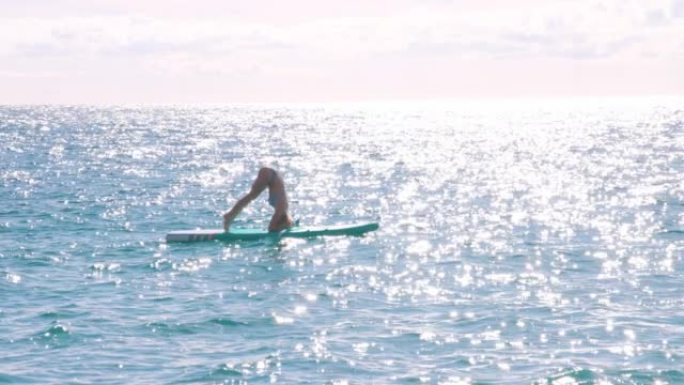 年轻的女飞行员在清澈的海水中站立在站立的桨板 (SUP) 上进行倒立