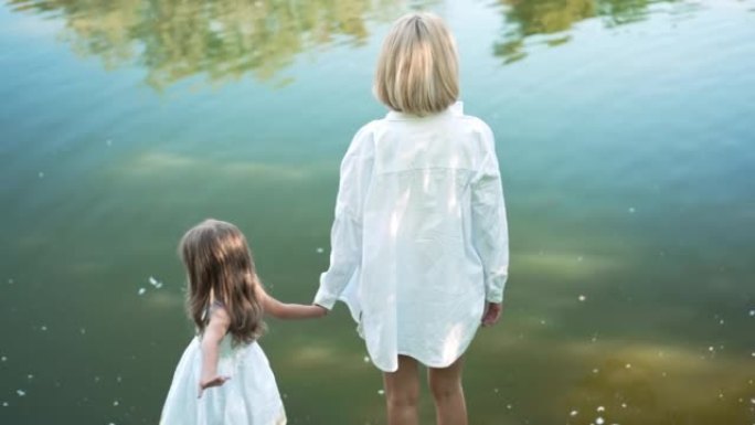幸福的母女手牵着手站在夏季春季公园的湖水背景下。后视图快乐的高加索女人和女孩在慢动作中聊天享受休闲。
