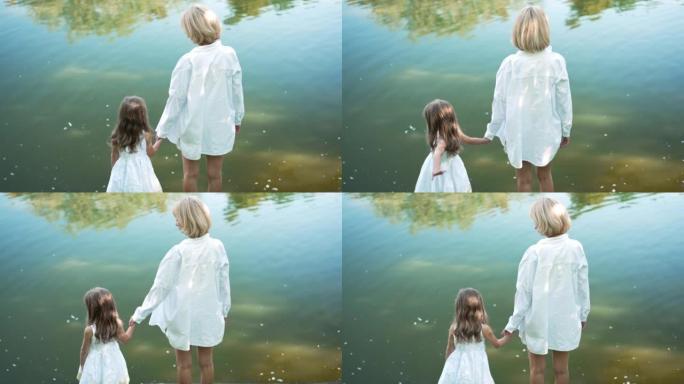 幸福的母女手牵着手站在夏季春季公园的湖水背景下。后视图快乐的高加索女人和女孩在慢动作中聊天享受休闲。