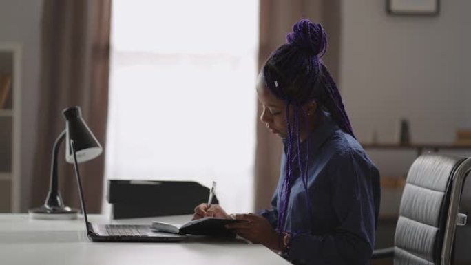 成年黑人女学生正在在线学习，在笔记本电脑屏幕上观看讲座，并在笔记本电脑上做笔记，坐在公寓房间的桌子上