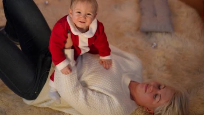 可爱的女婴笑的高角度视图，模糊的女人手里拿着孩子躺在家里的地毯上。穿着圣诞老人服装的快乐高加索蹒跚学