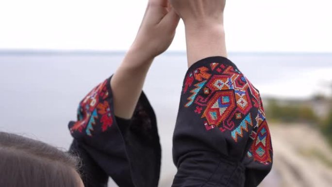 站在户外的难以辨认的年轻女子举手的黑色连衣裙上的特写传统刺绣。放松的乌克兰自信女士在河岸享受秋天的休
