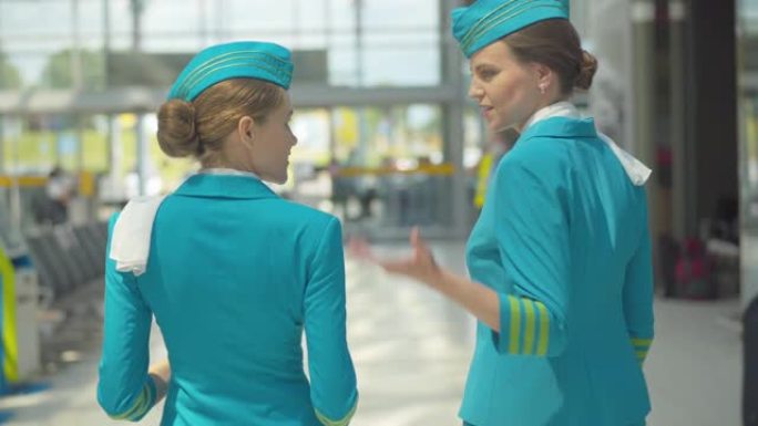 两名积极的空姐走到机场出口并交谈的后视图。摄像机跟随专业空姐回家。在航空工作的白人妇女的中景。