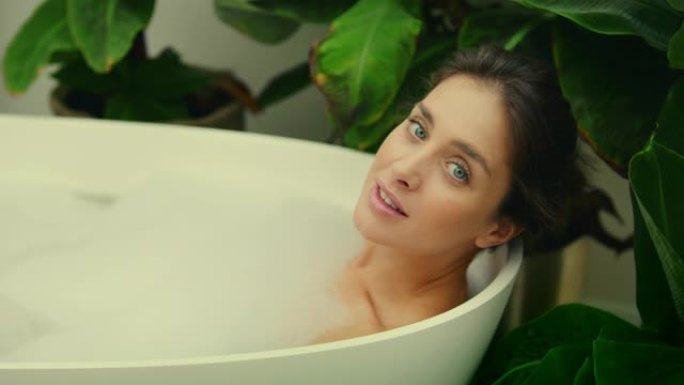 年轻女子在浴室内郁郁葱葱的绿色花园里洗泡泡浴。对着镜头微笑
