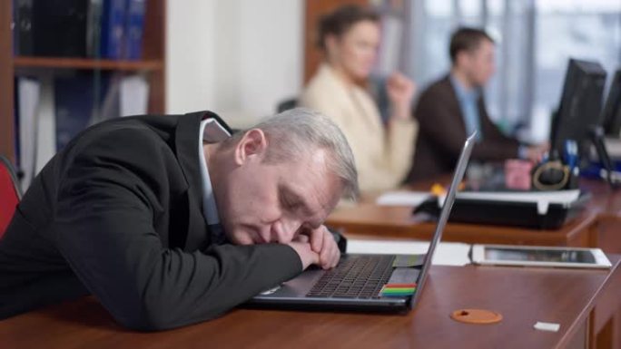 精疲力尽的高级男子睡在办公室的桌子上，模糊的同事在后台工作。疲惫不堪重负的白人男性员工在室内的肖像。