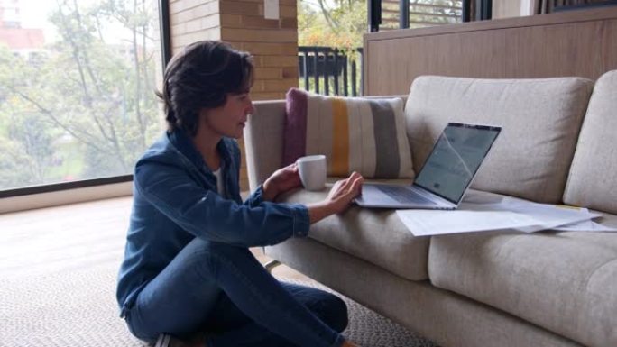 拉丁美洲放松的妇女在家工作，坐在地板上，用笔记本电脑一边喝咖啡一边看文书工作