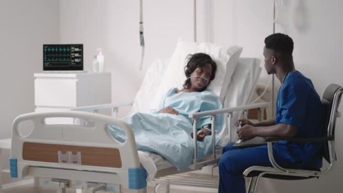 一名黑人男性医生正在与一名躺在医院病床上并与酸供应相连的黑人女性患者交谈。主治医师在医院与患者讨论治