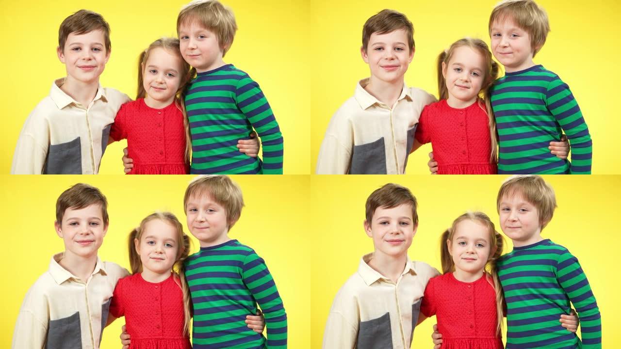 三个迷人的孩子在黄色背景下拥抱，微笑着看着相机。朋友或兄弟姐妹摆姿势的肖像。兄弟姐妹一起享受时光。团