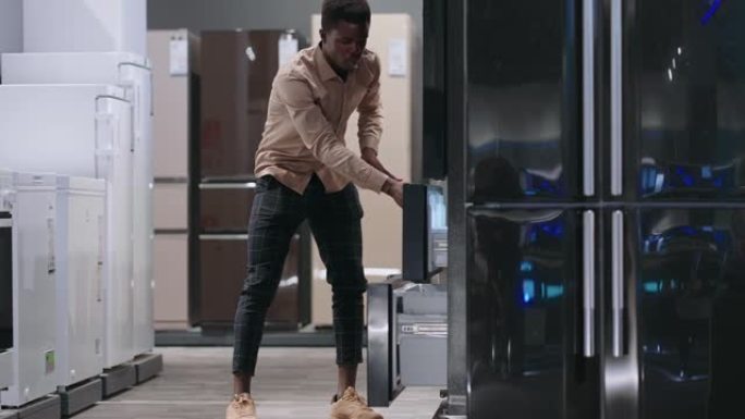 美国黑人男子在家电商店选择冰箱，在里面看展览样品，打开冰箱，全长肖像