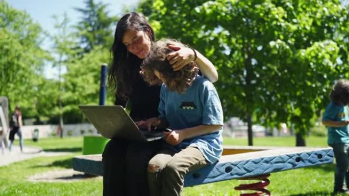 母亲与儿子一起在计算机上的户外长凳上工作