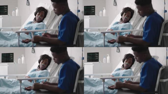 手术后在医院病床上休息的非裔美国妇女与年轻的男护士交谈。非裔美国护士协助躺在床上的女性患者的肖像