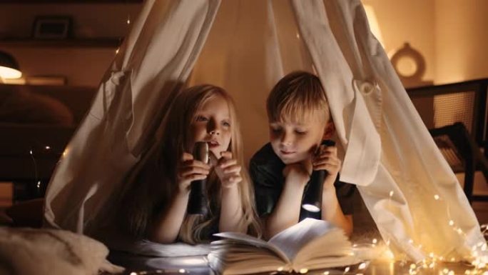 幸福的家庭。两个孩子女孩和男孩正在帐篷里看书。兄弟姐妹手中的手电筒。帐篷里有书的幸福家庭。孩子们坚持