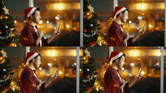 圣诞之夜，英俊的男人戴着圣诞帽，一边享受着热饮，一边玩智能手机，一边站在公寓的窗边。