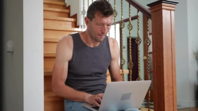男人坐在客厅的楼梯上喃喃地在笔记本电脑键盘上打字。成人高加索人的肖像在室内在家网上冲浪。生活方式和现