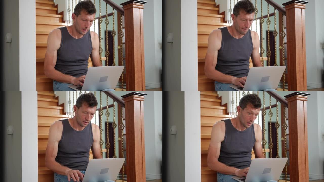 男人坐在客厅的楼梯上喃喃地在笔记本电脑键盘上打字。成人高加索人的肖像在室内在家网上冲浪。生活方式和现