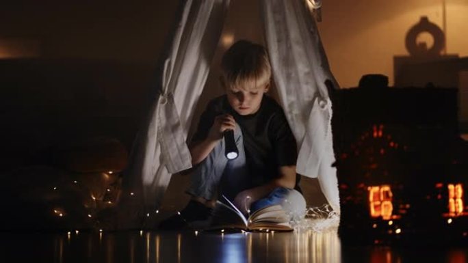 一个可爱的金发男孩在花环和灯的灯光下坐在黑暗中的小屋中的灯笼的灯光下看书。