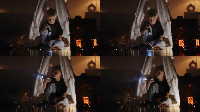 一个可爱的金发男孩在花环和灯的灯光下坐在黑暗中的小屋中的灯笼的灯光下看书。