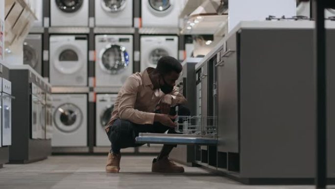 戴着口罩的非洲裔美国人在家电商店选择洗碗机，开门向内看