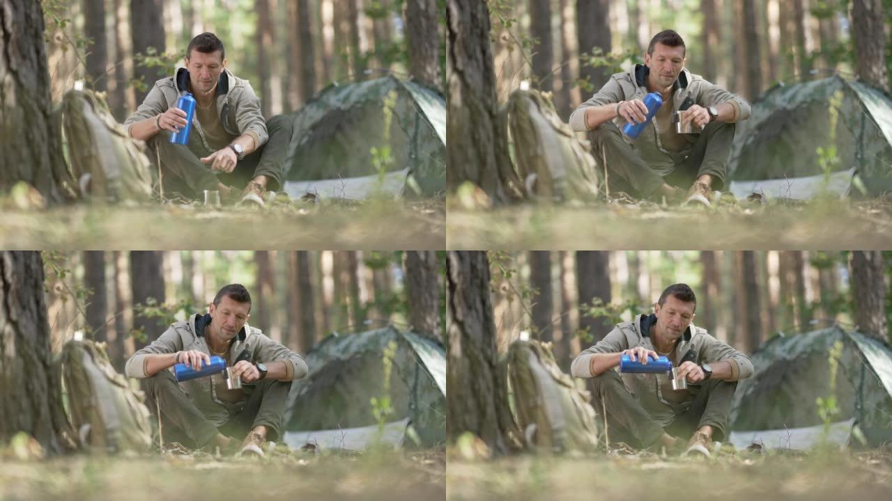 前视图快乐的男性游客坐在森林的露营地，慢动作地从热水瓶里倒饮料。夏天春天，宽镜头的白人在户外的木头上