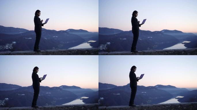 商人站在日落时俯瞰山脉和湖泊的壁架上，使用笔记本电脑