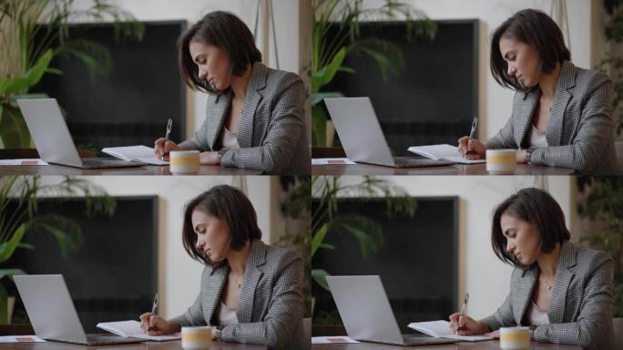 女商人分析图表并在笔记本电脑上打字。远程工作，戴眼镜的年轻女性在剪贴板上写笔记，在笔记本电脑键盘上打