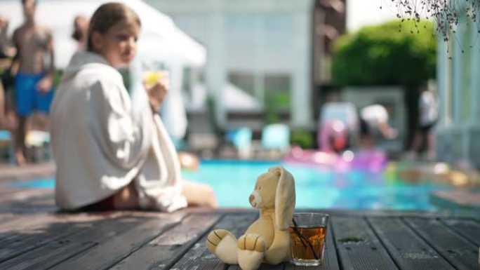 游泳池边甲板上健康的维生素汁和兔子玩具，模糊的女孩坐在毛巾的背景下喝酒。健康的生活方式和快乐的暑假理