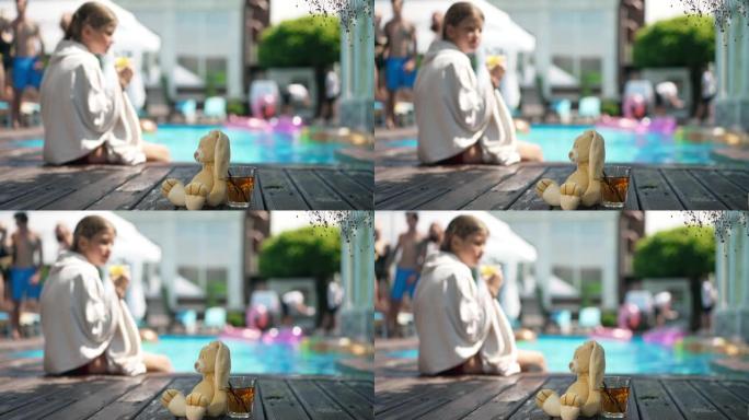 游泳池边甲板上健康的维生素汁和兔子玩具，模糊的女孩坐在毛巾的背景下喝酒。健康的生活方式和快乐的暑假理