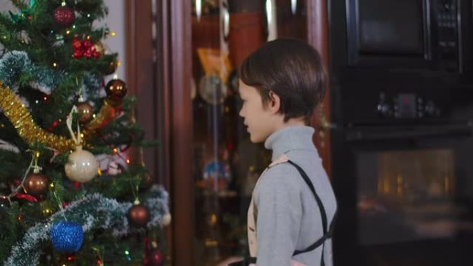 可爱的积极的高加索男孩走进房间，和圣诞树说话。积极的孩子在除夕夜向圣诞老人说祝福。假期和希望概念。电