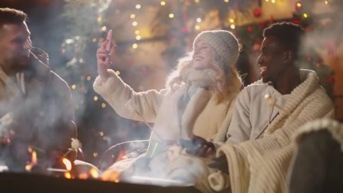 冬天圣诞节晚上，美丽的金发女人在篝火旁用智能手机自拍