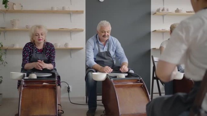 中年陶瓷艺术家教学小组老年白人妇女和高级男子如何在艺术工作室的办公桌上楔入粘土。人们喜欢在工作中交谈