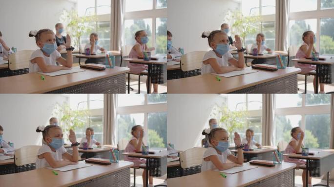学校教室里戴着面具的孩子坐在教室里，以慢动作举手回答老师的问题。在学校大流行期间的课程